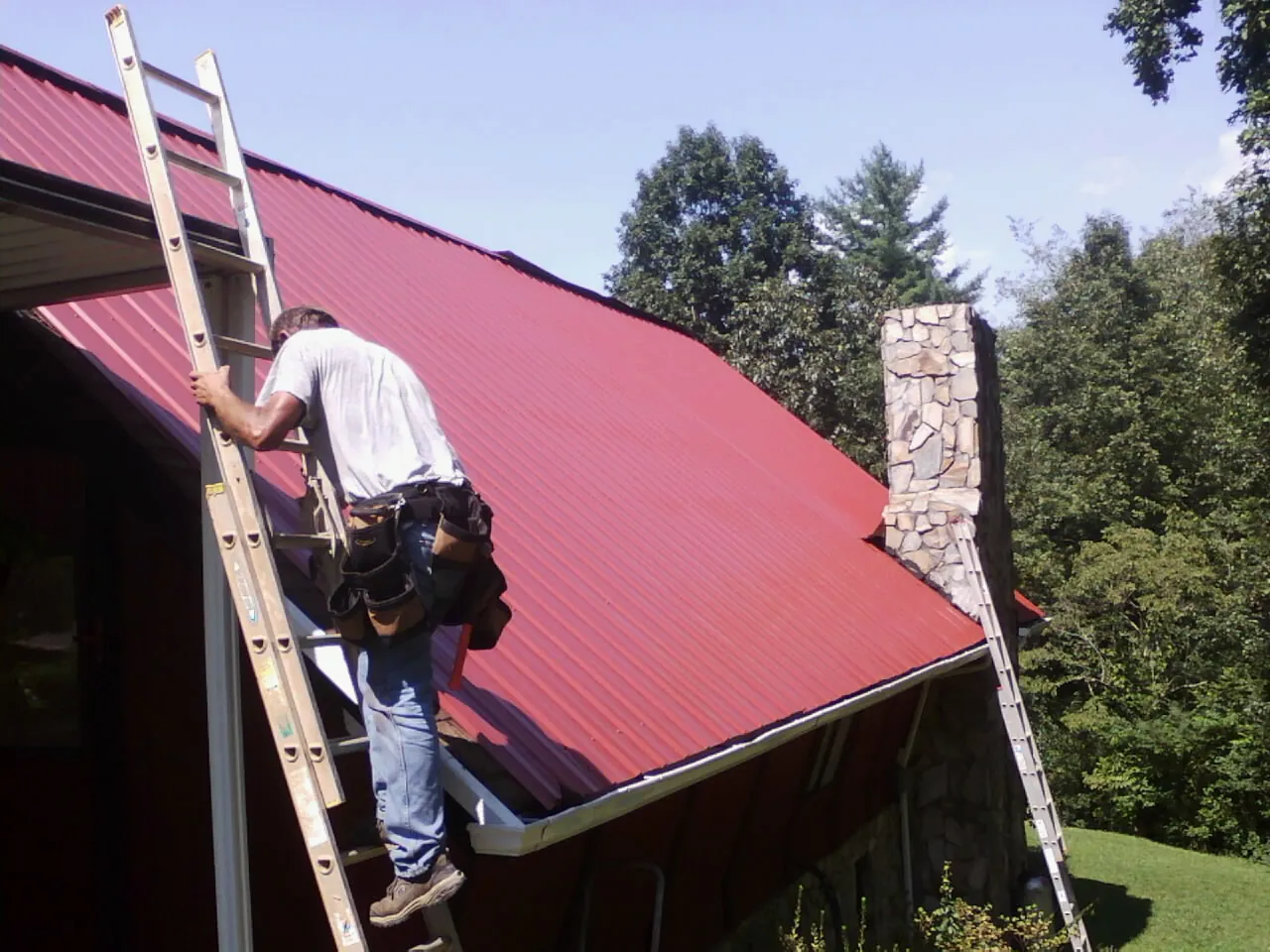Roofer on a ladder beside a red metal roof and stone chimney — Soffit & Fascia Repair by Roofing Brothers in Asheville